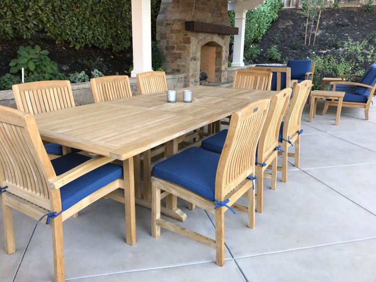 Summer Cedar An outdoor dining area featuring a large wooden table with matching chairs, blue seat cushions, set on a patio with a fireplace in the background, ready for a gathering or meal. Available at Summer Cedar. summercedar.com