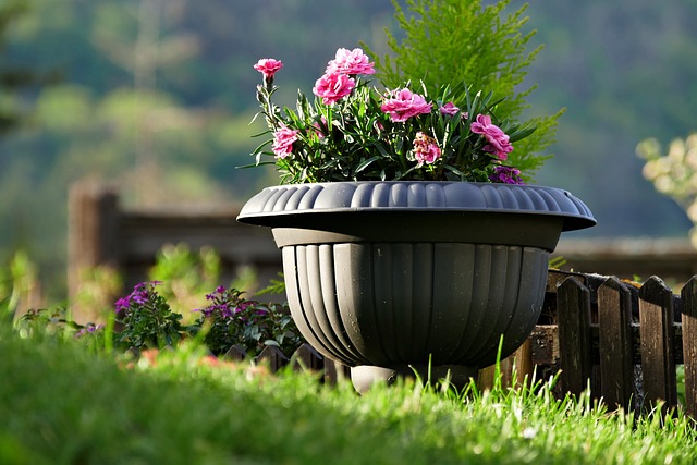 Summer Cedar A vibrant planter maximizing green spaces with blooming pink flowers and lush green foliage, positioned against a soft-focus natural backdrop, perched atop a neatly trimmed lawn beside a rustic wooden fence. summercedar.com