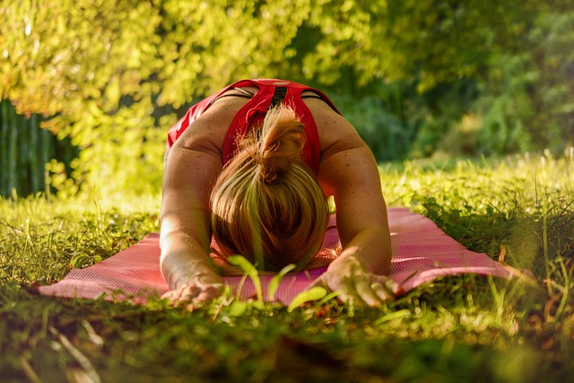Summer Cedar A woman practicing outdoor yoga on a pink mat in a serene, sunlit green park, performing a forward bend stretch that reflects tranquility and connection with nature. summercedar.com